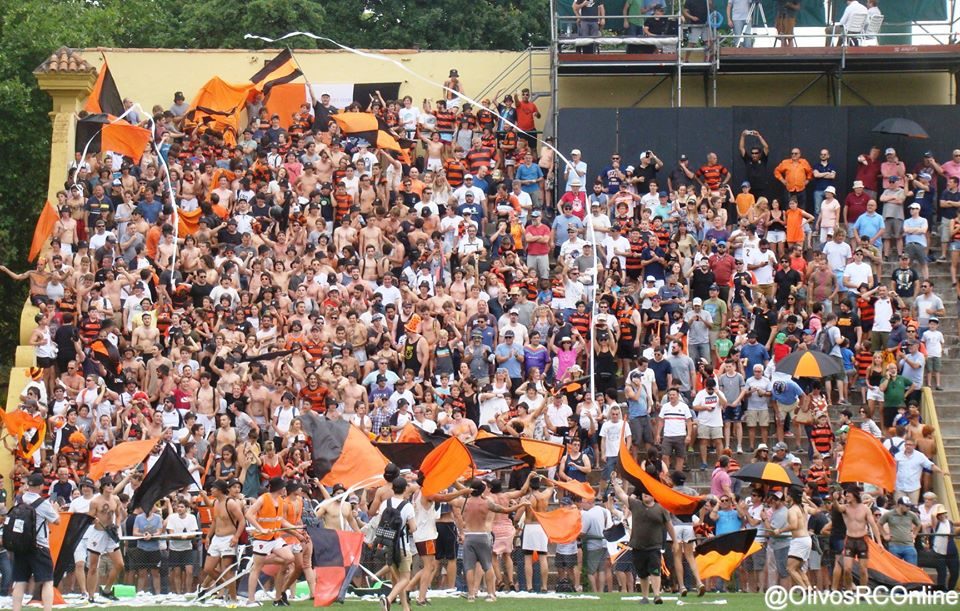 Hinchada de Olivos en la tribuna del club con banderas naranjas