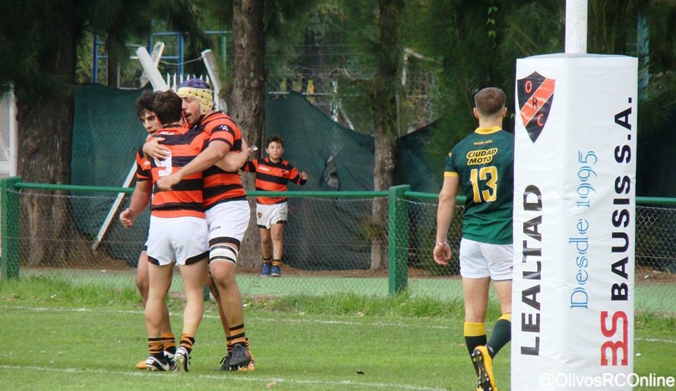 Dos jugadores de Olivos abrazados junto al poste con el escudo del club